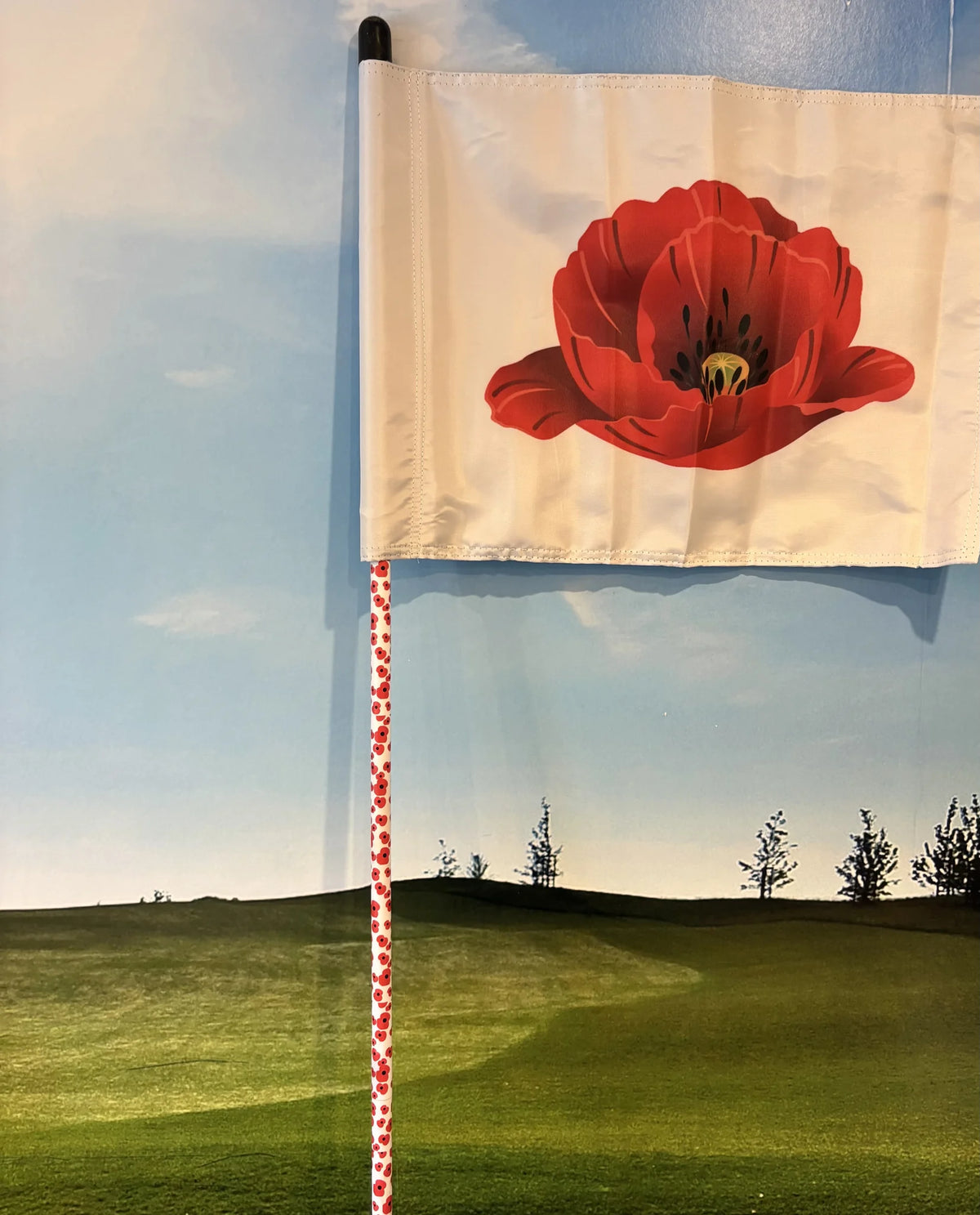 White tube lock golf flag printed with a single red poppy, flying against a sky and fairway backdrop